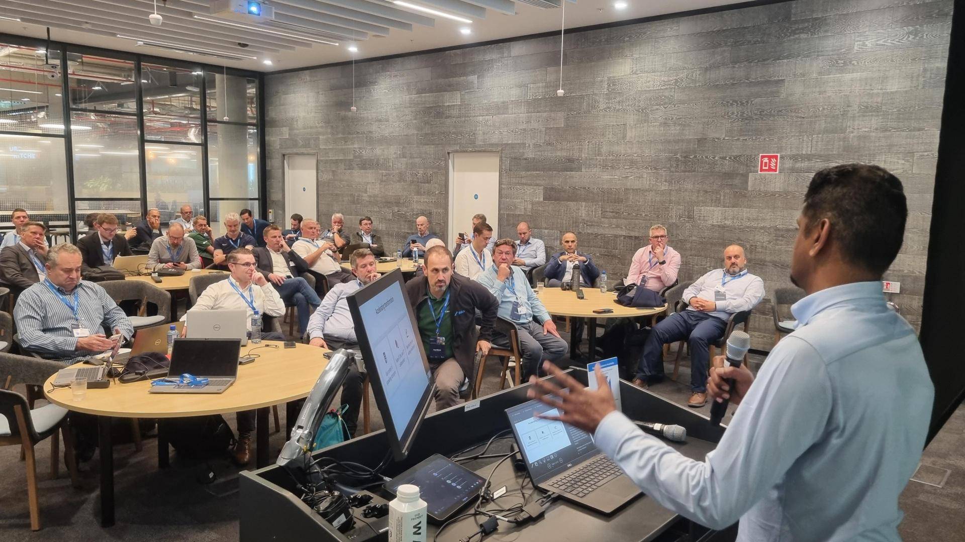Palanikumar, holding a microphone, presents to attendees seated at round tables with laptops.